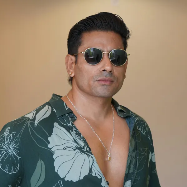 A stylish modern man with dense hair volume wearing round silver-framed sunglasses and an unbuttoned green floral shirt, displaying an invisible artisan custom hairline and premium professional men's hairpieces.