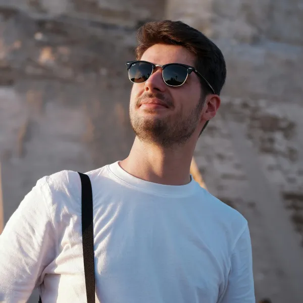 A sophisticated professional man with full, flawless hair volume wearing silver-framed aviator sunglasses, a white turtleneck, and a beige belted safari jacket, representing non-surgical hair replacement as the elite transplant alternative.